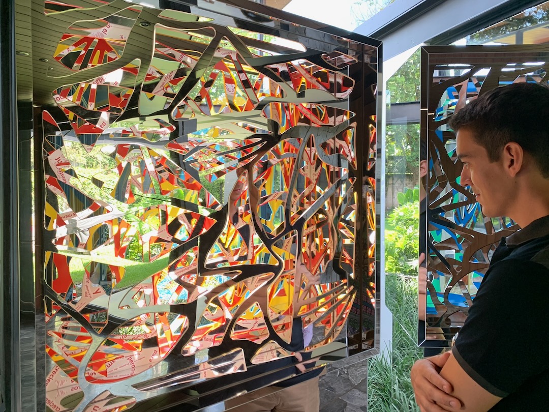 A visitor contemplating an Ambrosia panel painted in reds and yellows with words woven through, in a glass pavilion with a garden beyond