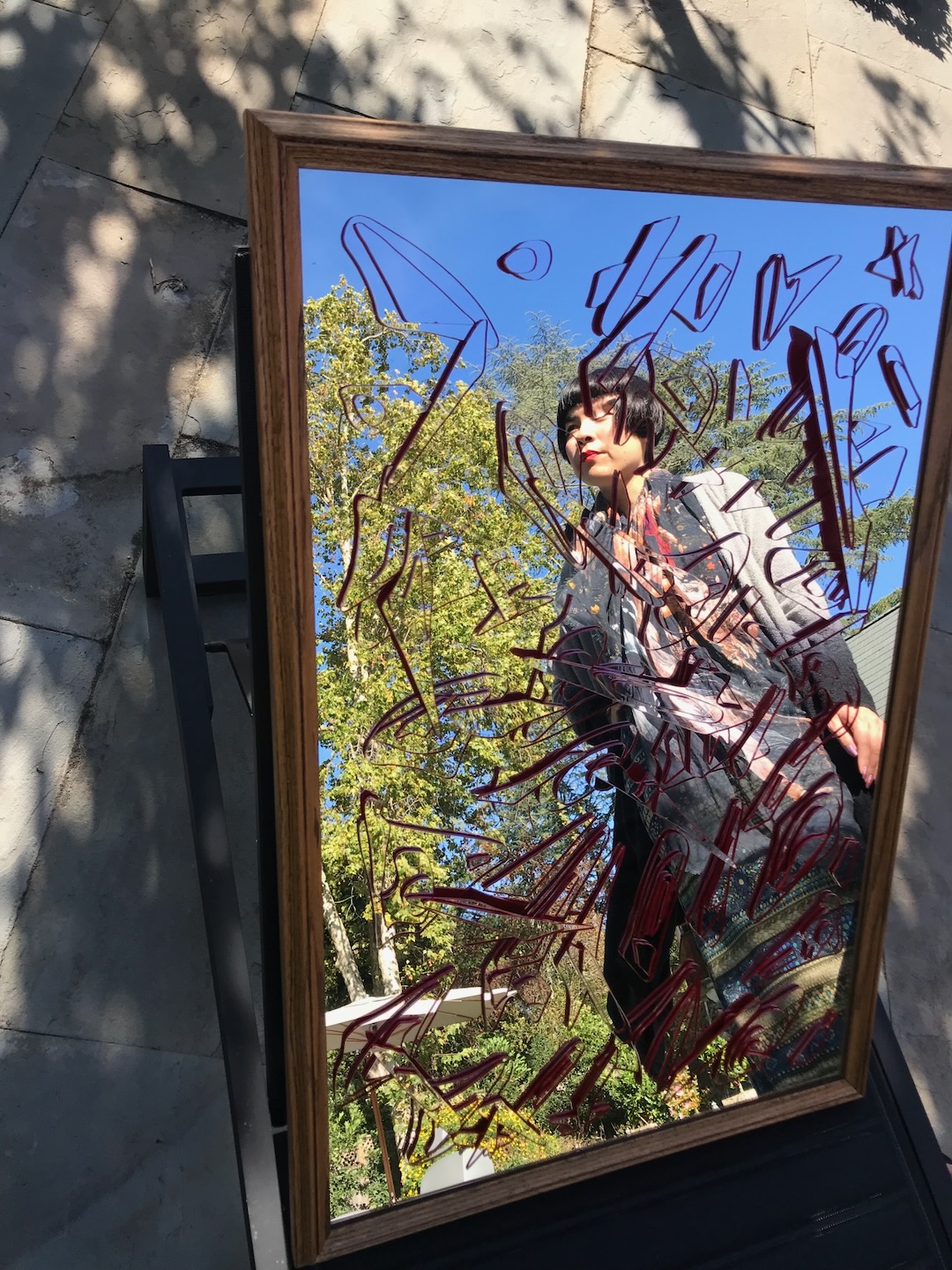 A framed Cloud Island panel on a stone terrace, its red-edged mirrored clouds reflecting Drue Kataoka, blue sky and garden trees