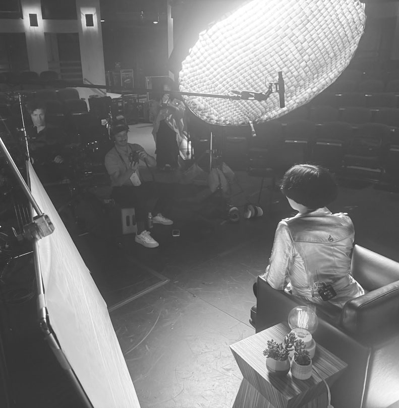 Drue Kataoka seated for a filmed interview on a darkened theater stage, a large softbox lighting the set as the crew films